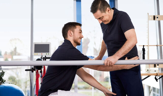 Therapeut übt mit Patient das Gehen an der Stange.